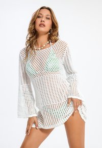 Bílý pletený plážový přehoz volného střihu s dlouhými zvonovými rukávy a texturovaným otevřeným vzorem. Má detaily vlnkovaného okraje.