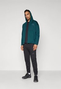 Camisola com capuz azul-petróleo, fecho éclair frontal, bolsos frontais e detalhes com logótipo, combinada com calças pretas e ténis. Tecido suave e corte descontraído.