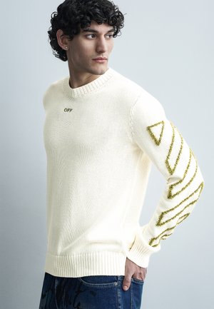Νέος άνδρας με σγουρά μαλλιά που φορεί λευκό πουλόβερ με πράσινα γεωμετρικά σχέδια στο μανίκι και το κείμενο «OFF» στο στήθος, κοιτάζει πλάγια.