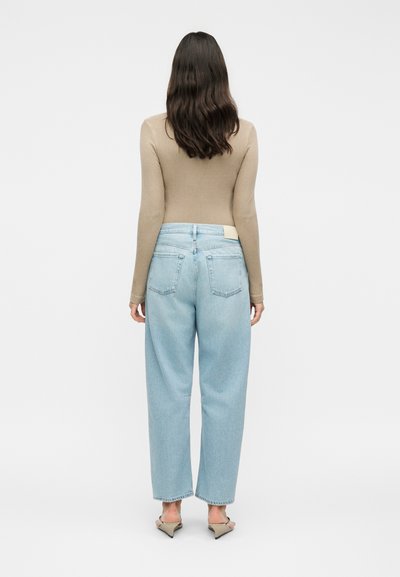 Γυναίκα με μακριά καστανά μαλλιά που φοράει μπεζ ριπ πουλόβερ, ανοιχτομπλε τζιν με ψηλή μέση και φαρδιά πόδια και μπεζ παπούτσια με χαμηλό τακούνι, στέκεται γυρισμένη προς τα πίσω.