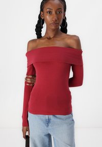 Raudonas off-the-shoulder viršus su ilgomis rankovėmis, prigludęs dizainas, minkšta tekstūra, be siūlių iškirptė, dėvimas su šviesiai mėlynomis džinsomis. Paprastas ir universalus.