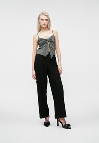 Γυναίκα που φοράει μια γκρίζα αμάνικη μπλούζα με κόμπο μπροστά, μαύρο παντελόνι σε φαρδιά γραμμή και μαύρες ψηλές γόβες με φιόγκο, που στέκεται μπροστά σε άσπρο φόντο.