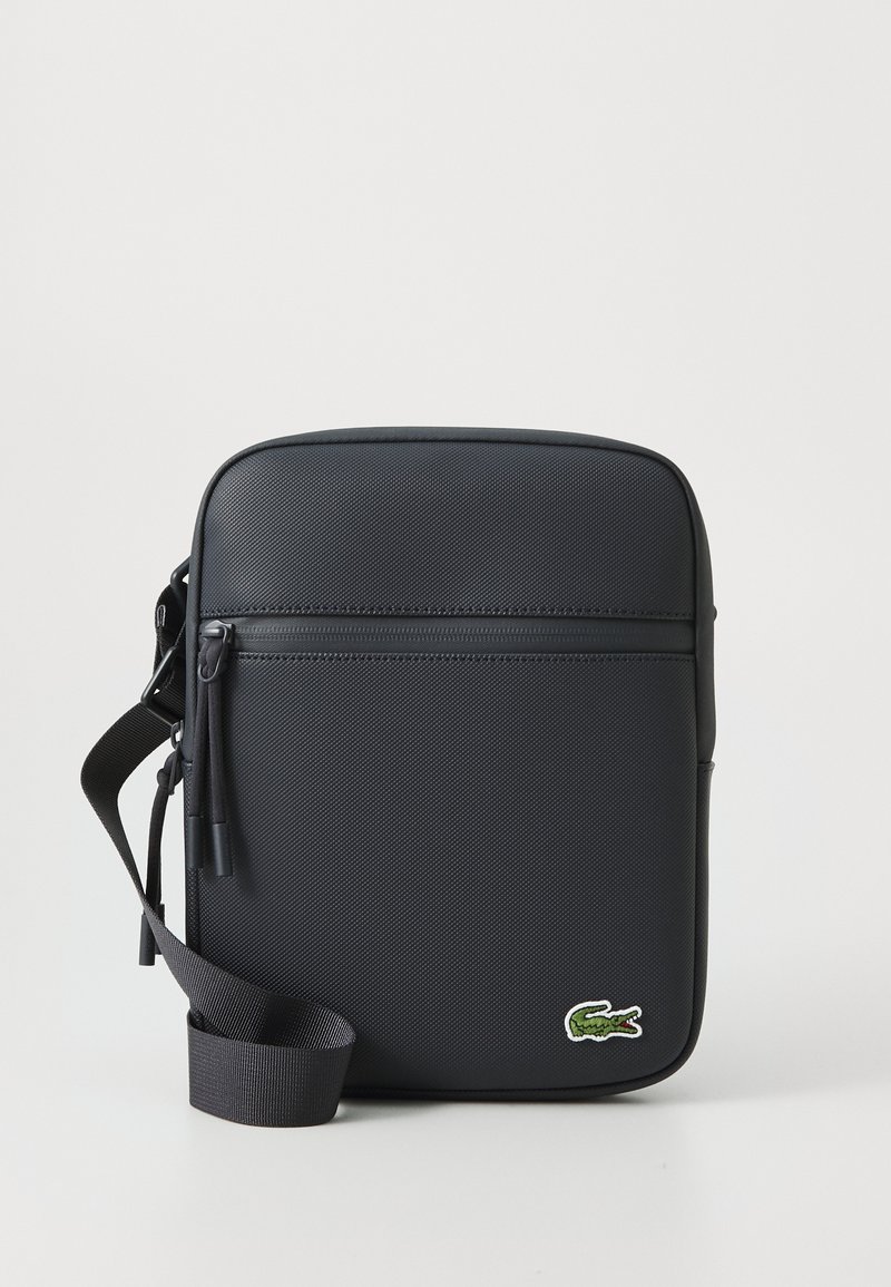Carteira cruzada preta com material texturizado, forma retangular, bolso frontal com fecho a zip e detalhe em logo de crocodilo verde. Inclui uma alça ajustável.
