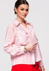 Femme portant un chemisier rose clair avec un grand nœud à l’avant et une jupe rouge avec un nœud sur le côté, accessoirisée de boucles d’oreilles créoles dorées et d’un bracelet.