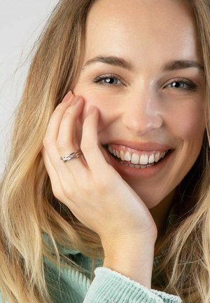 Bague en argent à motif tordu, portée sur une main touchant le visage. L'arrière-plan est neutre et la personne a les cheveux blonds mi-longs.