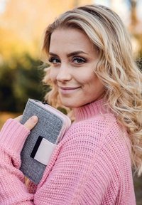 Gray textured wallet with a pink zipper and strap, held against a pink knitted sweater. Background features soft outdoor colors.