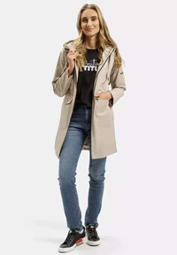 Beżowa wodoodporna kurtka z kapturem, z dwoma przednimi kieszeniami i zapięciem na suwak. Noszona na czarnej koszulce z nadrukiem oraz niebieskich dżinsach.