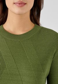 Pull en maille verte à côtes, avec un col rond et des détails texturés en diagonale. Le tissu possède une apparence douce et texturée.