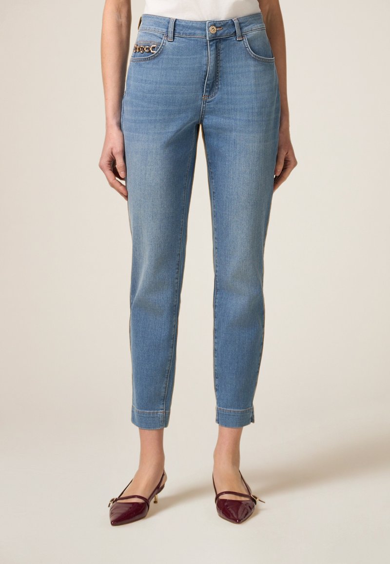 Jeans de mezclilla azul claro con un corte ajustado, que presentan una cadena decorativa dorada en el bolsillo izquierdo y los puños doblados en el tobillo.