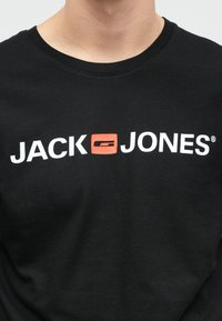 Černé bavlněné tričko s výrazným bílým textem "JACK" a "JONES" s červeným akcentním boxem mezi nimi. Design s kulatým výstřihem.