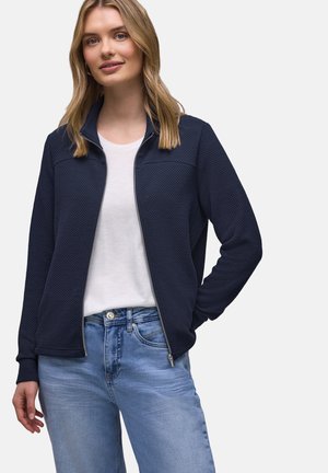 Kvinde iført en marineblå, tekstureret jakke med lynlås over en hvid t-shirt og lyseblå jeans, stående med den ene hånd i lommen.