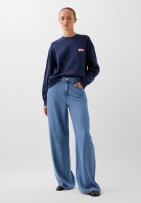 Felpa navy con logo rosa e vestibilità relaxed, abbinata a jeans in denim chiaro a gamba larga. Scarpe nere completano l'outfit.