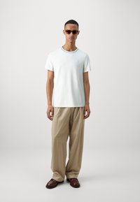 T-shirt a maniche corte verde chiaro con bordo nero, abbinato a pantaloni beige a gamba larga e scarpe marroni, con un piccolo logo sulla maglietta.