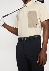 Polo beige avec une poche plaquée marron, une ceinture tressée noire et un pantalon sombre. Un club de golf dans une main, un gant dans l'autre.