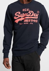 Sudadera azul marino hecha de tela suave, con un prominente logo y texto rosa "SuperDry" en el pecho, puños y dobladillo acanalados.