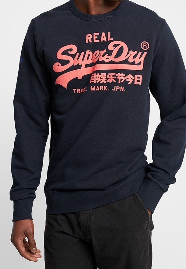 Sudadera azul marino hecha de tela suave, con un prominente logo y texto rosa "SuperDry" en el pecho, puños y dobladillo acanalados.
