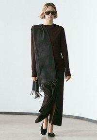 Brązowa bluzka z długim rękawem, ciemnoszary szal z frędzlami, zwiewna ciemna spódnica z wysokim rozcięciem, czarne płaskie buty oraz duże okulary przeciwsłoneczne.