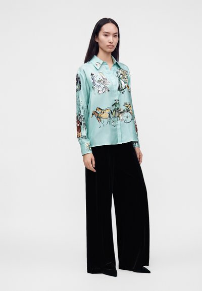 Femme debout portant une chemise en soie turquoise avec des imprimés de calèches et un pantalon large noir, sur un fond blanc uni.