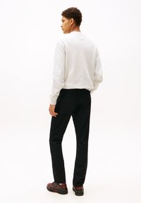 Młody mężczyzna stojący tyłem, ubrany w jasnoszarą bluzę, czarne spodnie i czarne czerwone trampki na białym tle.