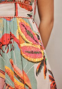 Gros plan d’une robe avec des motifs colorés de fruits tropicaux et de crabes, présentant des nuances orange, rose, vertes et jaunes avec un travail décoratif de perles.