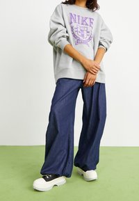 Szary, oversize'owy sweatshirt z fioletowym logo Nike, zestawiony z luźnymi, ciemnoniebieskimi spodniami dżinsowymi i masywnymi, białymi trampkami na zielonej powierzchni.
