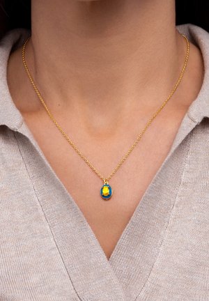 Collier chaîne en or avec pendentif en pierre précieuse ovale bleue et jaune porté autour du cou d'une personne, au-dessus d'un pull beige à col en V.