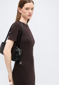 Femme portant une robe marron ajustée à manches courtes, tenant un petit sac à bandoulière noir avec une fermeture éclair en forme de cœur.