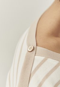 Cardigan rayé avec des bandes beige et crème, doté d'une texture lisse et d'un détail de bouton doré près de l'encolure.