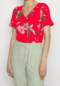 Blusa floreale rossa con scollatura a V e maniche corte, con fiori rosa e foglie verdi, abbinata a pantaloni leggeri verde chiaro con coulisse.