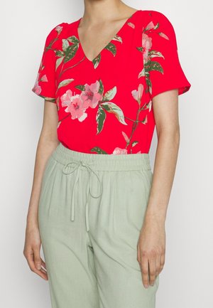 Rød blomsterbluse med V-hals og korte ærmer, prydet med pink blomster og grønne blade, parret med lys grønne bukser med snore.