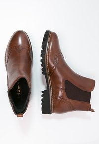 Bottines Chelsea en cuir marron avec des détails en brogue, panneaux latéraux élastiques et semelles crantées noires, présentées en vues de dessus et de côté.