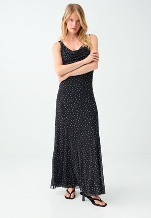 Femme blonde portant une robe maxi noire sans manches à pois et des sandales noires à talons, debout bras croisés.