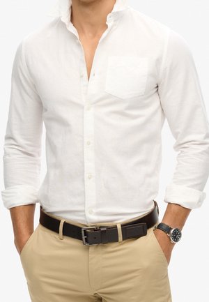 Homme portant une chemise blanche boutonnée avec les manches retroussées, un pantalon beige, une ceinture noire et une montre-bracelet noire à cadran rond.