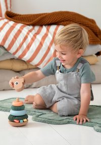 Jeune enfant assis par terre jouant avec un jouet empilable coloré en forme d’anneaux et de tête d’animal sur un tapis vert doux.