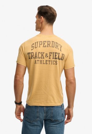 Hombre de espaldas vistiendo una camiseta amarilla Superdry Track & Field Athletics y vaqueros azules contra un fondo blanco liso.