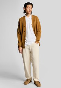 Brun cardigan over en hvid polo, cremede bukser og brune sko. Cardiganen har ribbede manchetter og frontknapper. Simpelt design.