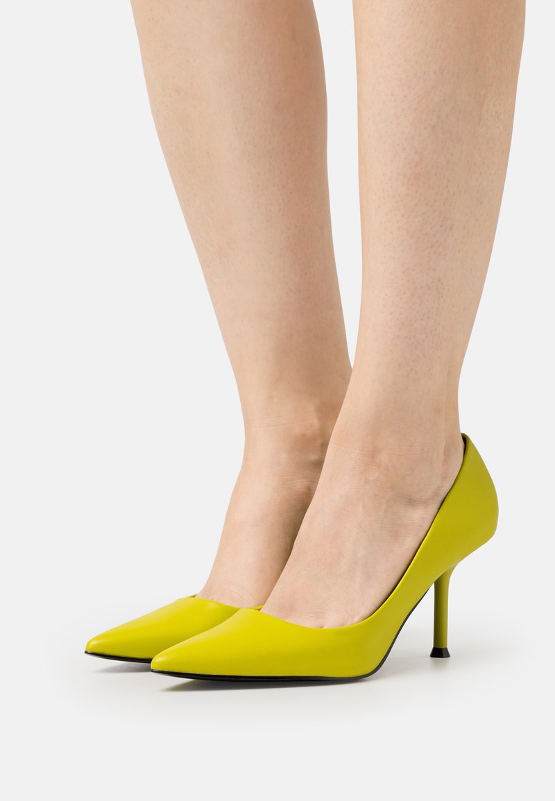 Jambes portant des escarpins pointus jaune vif à talons aiguilles fins sur un fond clair.