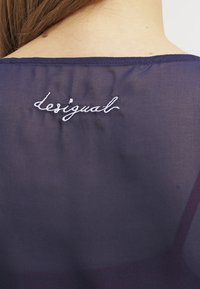 Dos d'une personne portant un haut transparent bleu foncé avec le logo brodé blanc "desigual" près de l'encolure.