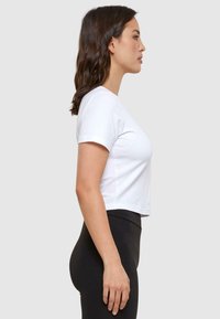 Witte cropped t-shirt met korte mouwen, gemaakt van zachte stof, gepresenteerd op een persoon die zwarte leggings draagt tegen een grijze achtergrond.