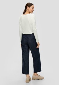 Langärmliges weißes Oberteil kombiniert mit dunkelblauen, weiten Hosen. Das Modell trägt beigefarbene Plateau-Schuhe. Glattes Material mit lässiger Silhouette.