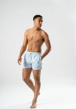 Muž bez košele stojaci bosý v svetlomodrých prúžkovaných šortkách s jednou rukou v vrecku, pozerajúci sa do strany na jednoduchom pozadí.