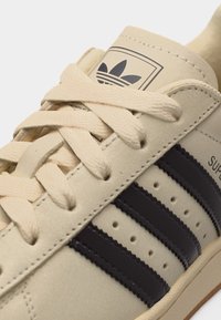 Μπεζ αθλητικό παπούτσι Adidas Superstar με μαύρες ρίγες, κρεμ κορδόνια και λογότυπο Adidas στη γλώσσα, εμφανίζοντας το πάνω μέρος και την περιοχή των δαχτύλων.