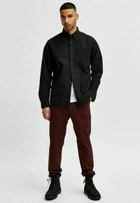 Homem de pé, vestindo uma camisa preta de botão, uma camisola branca por baixo, calças vermelhas escuras e botas pretas com cadarços, contra um fundo simples.