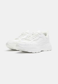 Scarpe da ginnastica bianche con una suola spessa, superiore strutturato e punta rotonda. Presentano lacci e contorni di design sottili sui lati.