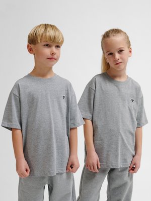 Dwoje dzieci stoi obok siebie, ubranych w pasujące szare, za duże t-shirty i spodnie z małym czarno-białym logo na piersi.