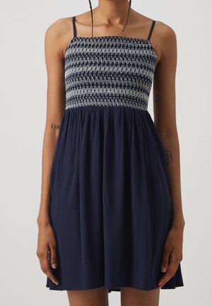 Marineblaues Kleid mit einem gerafften Oberteil und dünnen Trägern, das einen strukturierten, locker fallenden Rock aufweist. Keine auffälligen Muster oder Verzierungen.