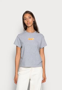 Szürke pamut póló rövid ujjal, kerek nyakkal, narancssárga "Levi's" logóval a mellkason. Világos színű nadrággal viselve.