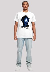 White cotton t-shirt featuring a blue graphic design of a face. Model wears light blue jeans and white sneakers.
