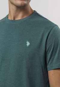 Grönblå t-shirt med korta ärmar, tillverkad av mjuk bomull, med en liten vit broderad logga på bröstet. Klassisk rund halsringning.