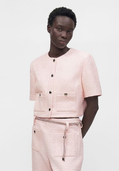 Femme portant une veste texturée rose clair à manches courtes et un pantalon taille haute assorti avec de gros boutons et une fine ceinture en corde.